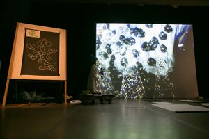 Dark art installation with chalkboard drawing, projected bubbles on screen, and shadowed figures in a gallery.