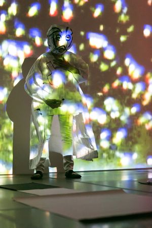 A figure in a white geometric costume and glowing mask stands before a colorful bokeh light projection backdrop.