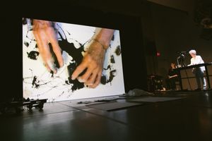 Artist performs live as large screen displays hands creating black ink art in a dark performance venue.