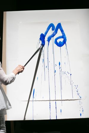 Artist painting bold blue abstract strokes on white canvas using a long-handled brush, with paint dripping down.