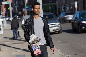 Young man in leather jacket carrying newspapers on a city street.