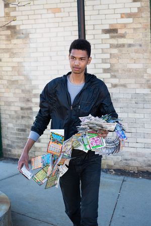Young man holding a large stack of lottery scratch tickets and newspapers on a city sidewalk.
