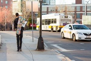 Person with colorful backpack standing at urban street corner with city bus and taxi cab in background.