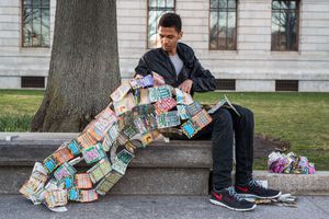 Man sitting outdoors on stone ledge examining a large quilt made of colorful lottery scratch tickets.
