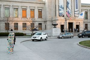 Person in colorful outfit stands outside a grand museum building with cars passing on a wide plaza.