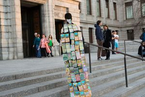 Person wearing colorful tiled coat art installation on museum steps with visitors in background.