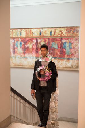 Young man holding a floral wreath stands on museum stairs before a colorful ancient fresco artwork.
