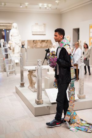 Man wearing colorful lottery ticket costume stands in classical sculpture museum gallery with ancient Greek statues.
