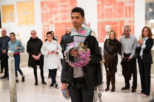 Young man holding colorful wreath artwork in gallery with crowd of visitors viewing exhibition behind him.