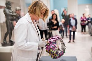 Museum conservator in white lab coat and gloves carefully examines colorful wreath artwork on display pedestal.