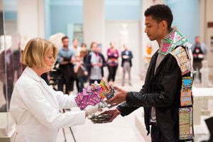 Woman in white coat hands colorful items to a young man in a decorated jacket at an indoor gallery event.