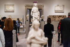 Visitors explore ancient Greek and Roman sculptures in a classical museum gallery with marble statues and decorative friezes.