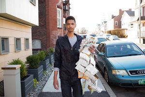 Young man carrying a large bundle of newspapers while standing on a residential street next to a teal Audi car.