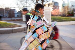 Person wearing a colorful outfit made of cereal boxes walking outdoors in an urban park setting.