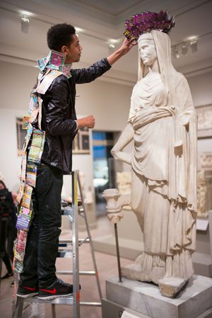 Artist on ladder decorating classical marble statue with colorful crown in museum gallery.