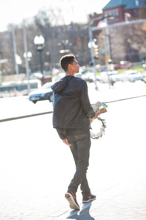 Young man in black jacket walking outdoors holding colorful items in a sunny urban setting.