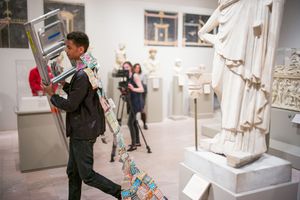 Person carrying artwork through a classical sculpture gallery museum exhibition with statues and visitors in background.