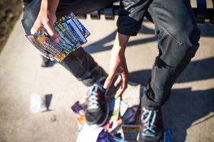 Person in dark pants holding colorful magazines near sneakers on a sunny sidewalk.