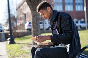 Young man in black jacket sitting on bench outdoors, reading documents on a sunny day.