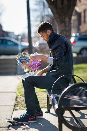 Young man sitting on a park bench sorting through colorful trading cards on a sunny day.