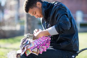 Young man sitting outdoors on a bench, scratching lottery tickets in a sunny park setting.