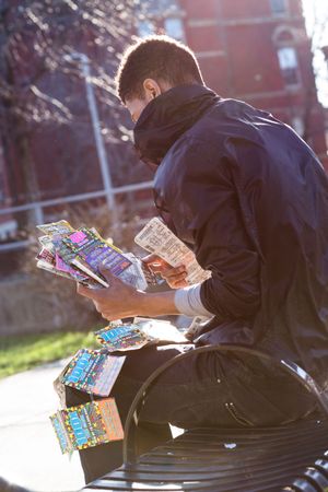 Man in dark jacket sits on bench examining colorful lottery scratch tickets outdoors on sunny day.