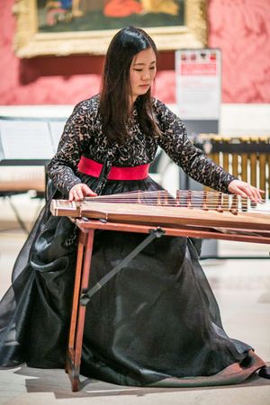 Woman in black dress with red sash playing a guzheng instrument indoors.