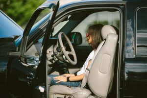 Person with sunglasses sitting in driver seat of black SUV with door open, using a laptop.