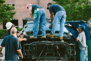 Workers in blue uniforms disassemble a large vehicle engine outdoors near a brick building.