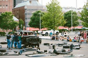 Dismantled car parts scattered in a parking lot with people gathering around a damaged vehicle at an outdoor urban event.