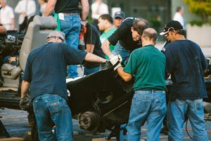 A group of mechanics in blue uniforms work together to move a race car component at an outdoor motorsport event.