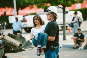 Man in hard hat and safety glasses carries woman on a city street near construction debris outdoors.