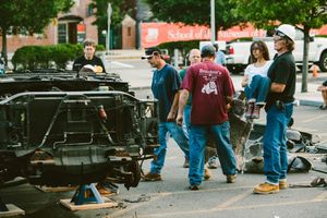 Workers in blue jeans examine disassembled vehicle parts on a city street near a School of Life building.