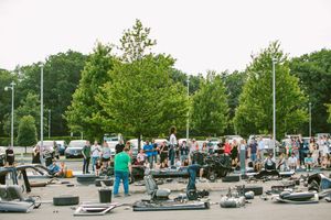 Crowd of people gathered in a parking lot surrounded by go-kart parts and tires with lush green trees in background.