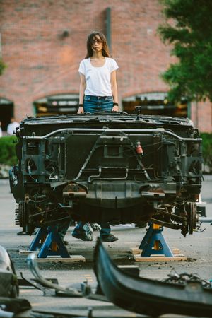 Young woman in white t-shirt and jeans standing on a disassembled car chassis supported by jack stands outdoors.