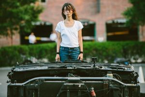 Young woman in white t-shirt and jeans standing behind an open car hood outdoors near a brick building.