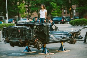 People examining a heavily damaged car chassis elevated on jack stands in a parking lot.