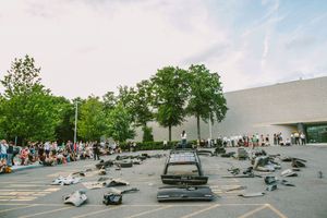 Crowd gathers outside modern building watching scattered car parts debris display in parking lot art installation.