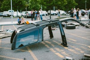 Overturned car wreck in parking lot with debris scattered around and bystanders observing the accident scene.