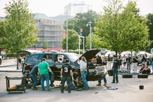 Mechanics and spectators gather around a black SUV with open hood in a sunny parking lot during an outdoor car event.