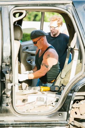 Two mechanics working on a vehicle interior, one tattooed man in cap and sunglasses, another in glasses observing repairs.