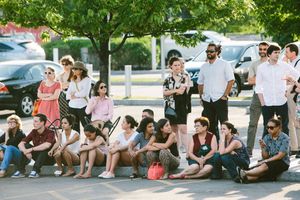 A diverse crowd of people sitting and standing outdoors on a sunny day near a parking lot with green trees.