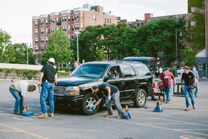 Group of people washing and cleaning a black GMC SUV in an urban parking lot with brick buildings in background.