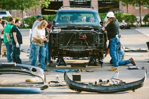 Group of mechanics disassembling a black SUV in a parking lot, with car parts scattered on the ground.