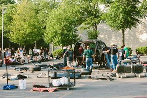 Mechanics and crew members work on vehicles in a busy outdoor parking lot surrounded by tires and tools.