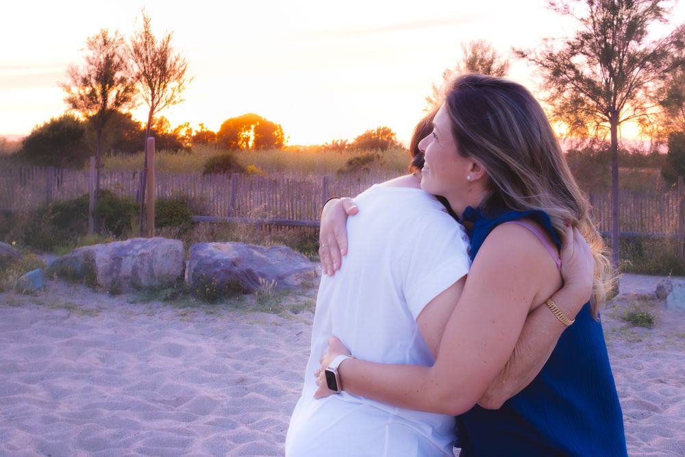 Two women embracing warmly at sunset in a sandy outdoor setting with trees and rocks in the background.