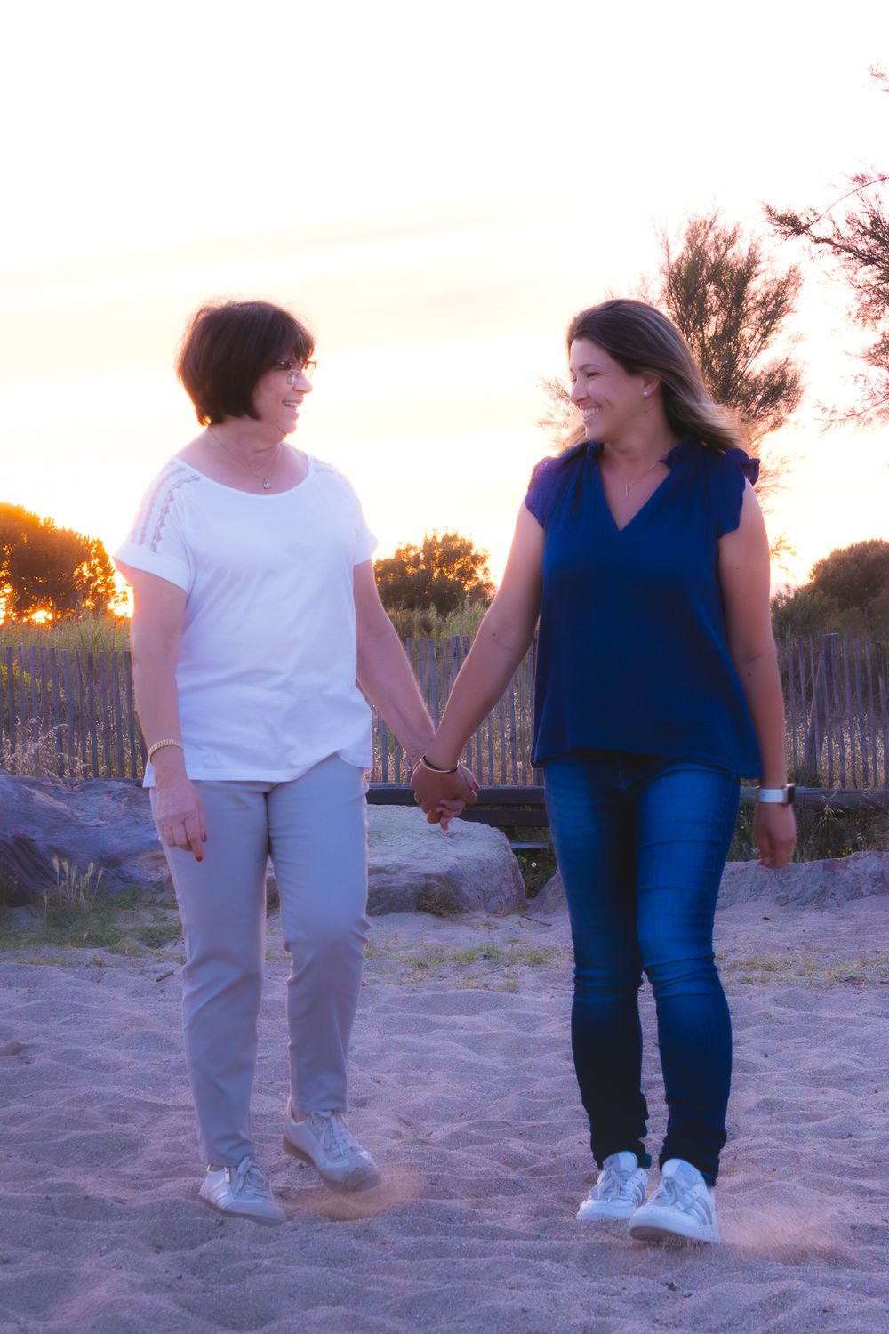 Two women holding hands and smiling at each other while walking on a sandy beach at sunset.