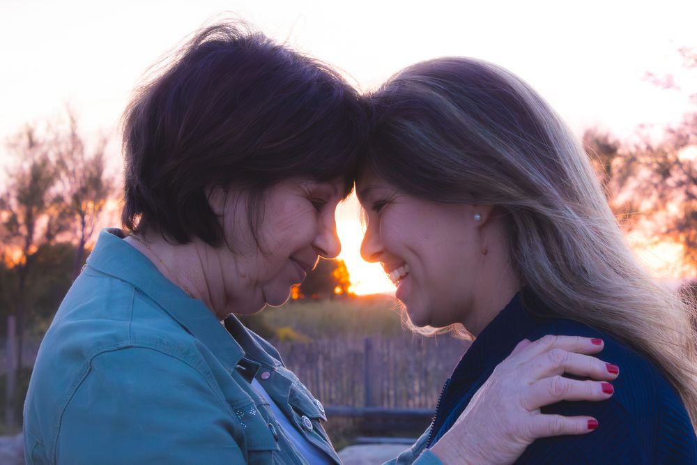 Two women share an intimate forehead-to-forehead moment at sunset, smiling warmly in a tender embrace outdoors.