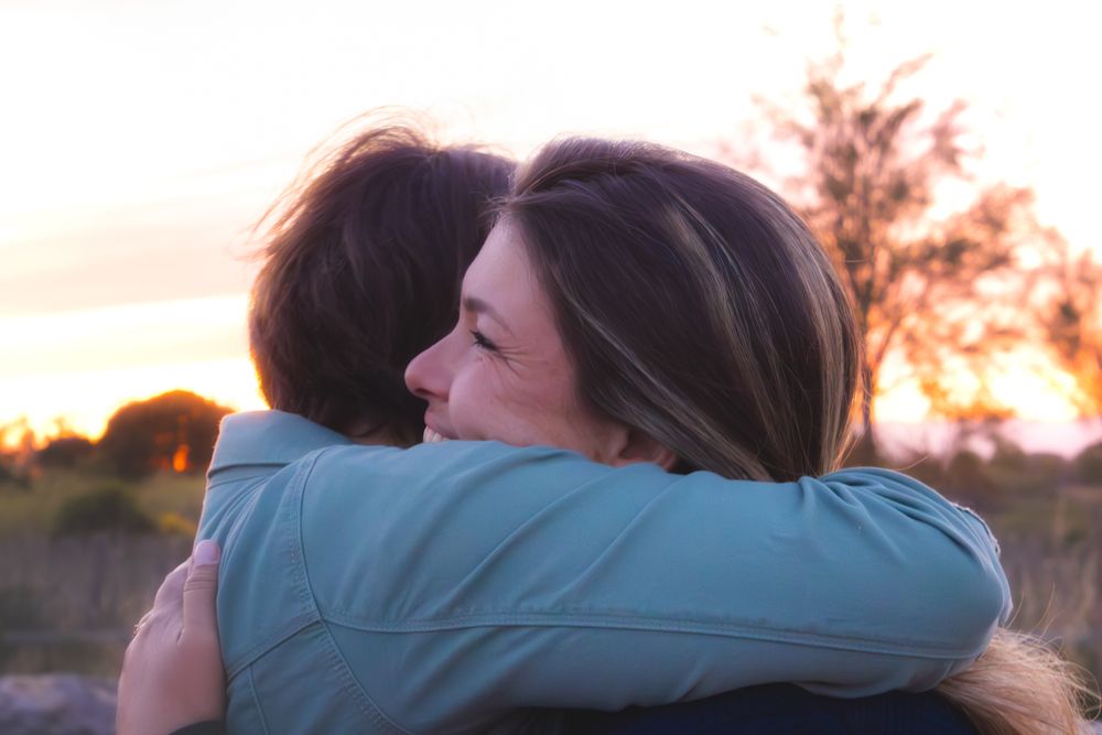 Two women sharing a warm embrace outdoors at sunset, with golden light illuminating their smiling faces.