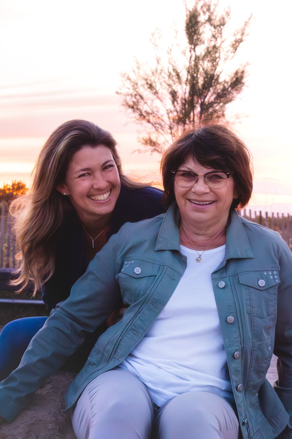 Two smiling women pose together outdoors at sunset, with trees and a colorful sky in the background.